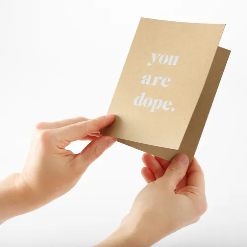 Two hands holding a custom card printed on Kraft paper with you are dope. in white ink on the front.