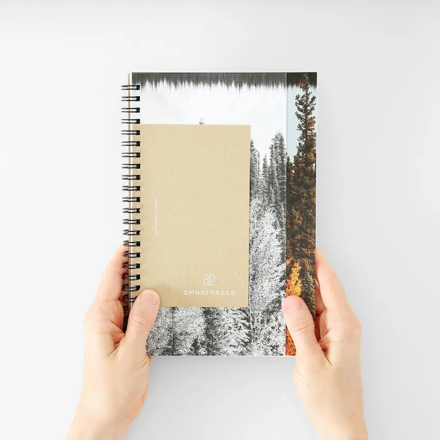 Two hands holding a custom notebook printed with a spiral binding and an outdoor scene with white ink on the cover.