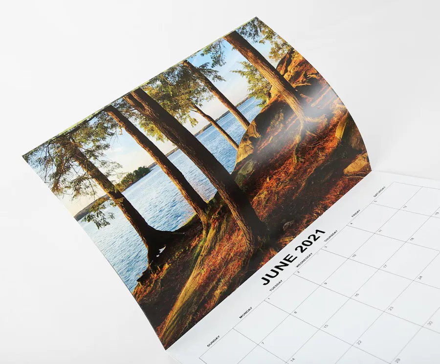 A custom wall calendar with a saddle stitch binding open to a nature scene of trees by a lake.