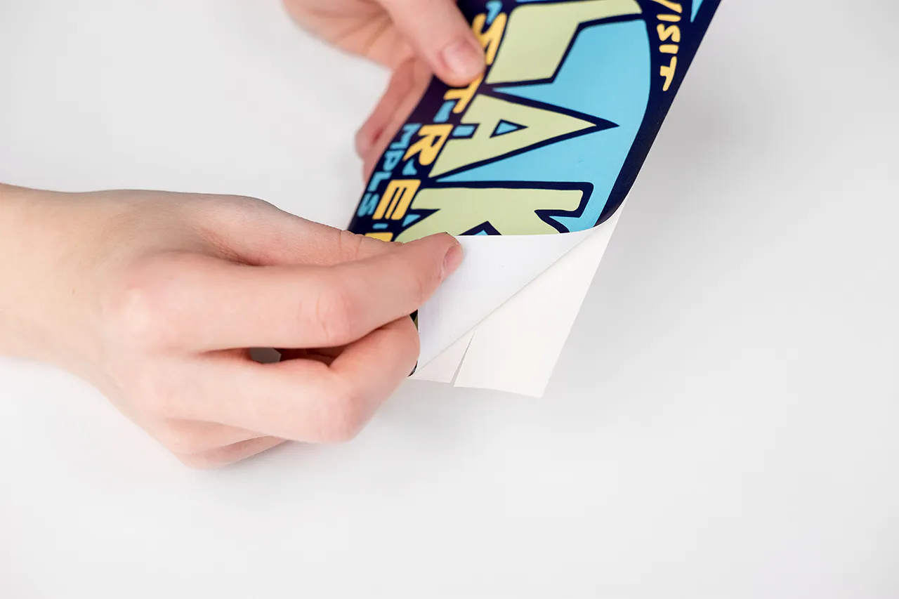 Two hands peeling a custom decal off its backing, printed with Visit Lake Street in blue, yellow and green.