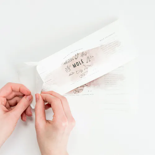 Two hands peeling a product label off its backing, printed with Mole and graphics of vegetables in the middle.