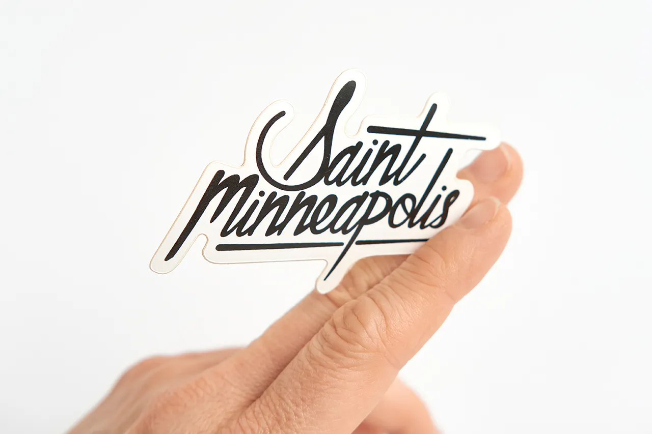 A custom die-cut sticker printed with Saint Minneapolis in black held between two fingers.