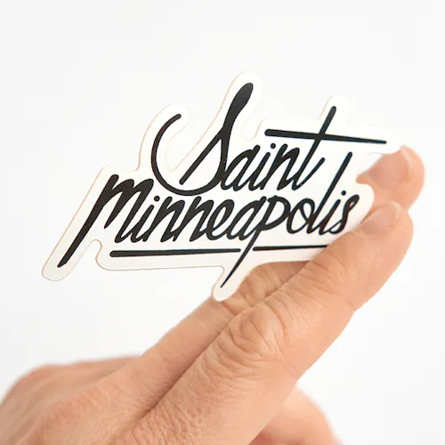 A custom die-cut sticker printed with Saint Minneapolis in black held between two fingers.