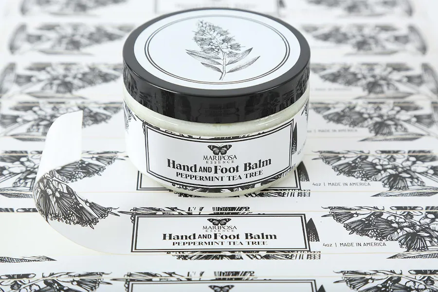 A jar with a custom product label printed with Hand and Foot Balm in black text sitting on a sheet of more labels.
