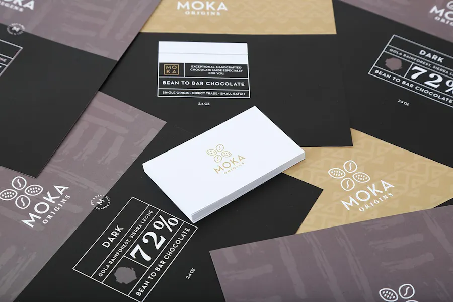 Moka Origins chocolate bar wrappers scattered below a stack of white business cards with Moka Origins in gold text.