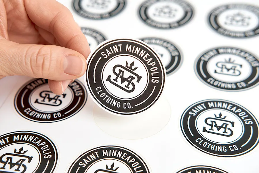A sheet of custom stickers printed with Saint Minneapolis Clothing Co. in white with a hand peeling one off.
