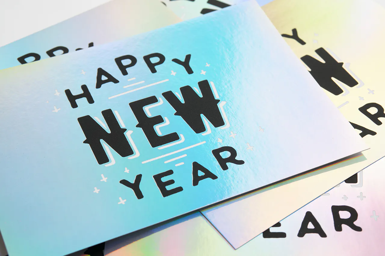 A pile of custom greeting cards printed with holographic foil and Happy New Year in black on the front.