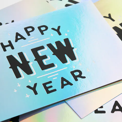 A pile of custom greeting cards printed with holographic foil and Happy New Year in black on the front.