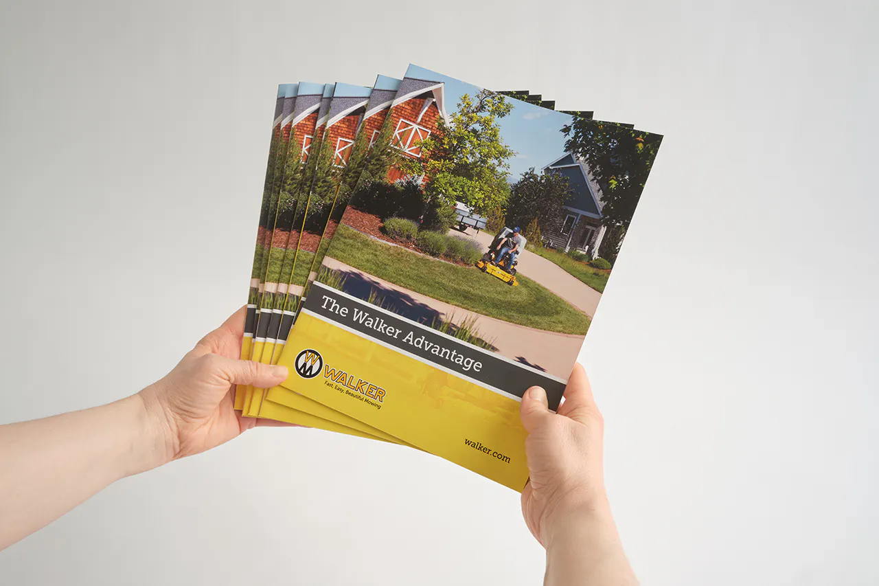 Two hands holding a stack of Walker Mower marketing manuals with a man on a riding mower on the cover.