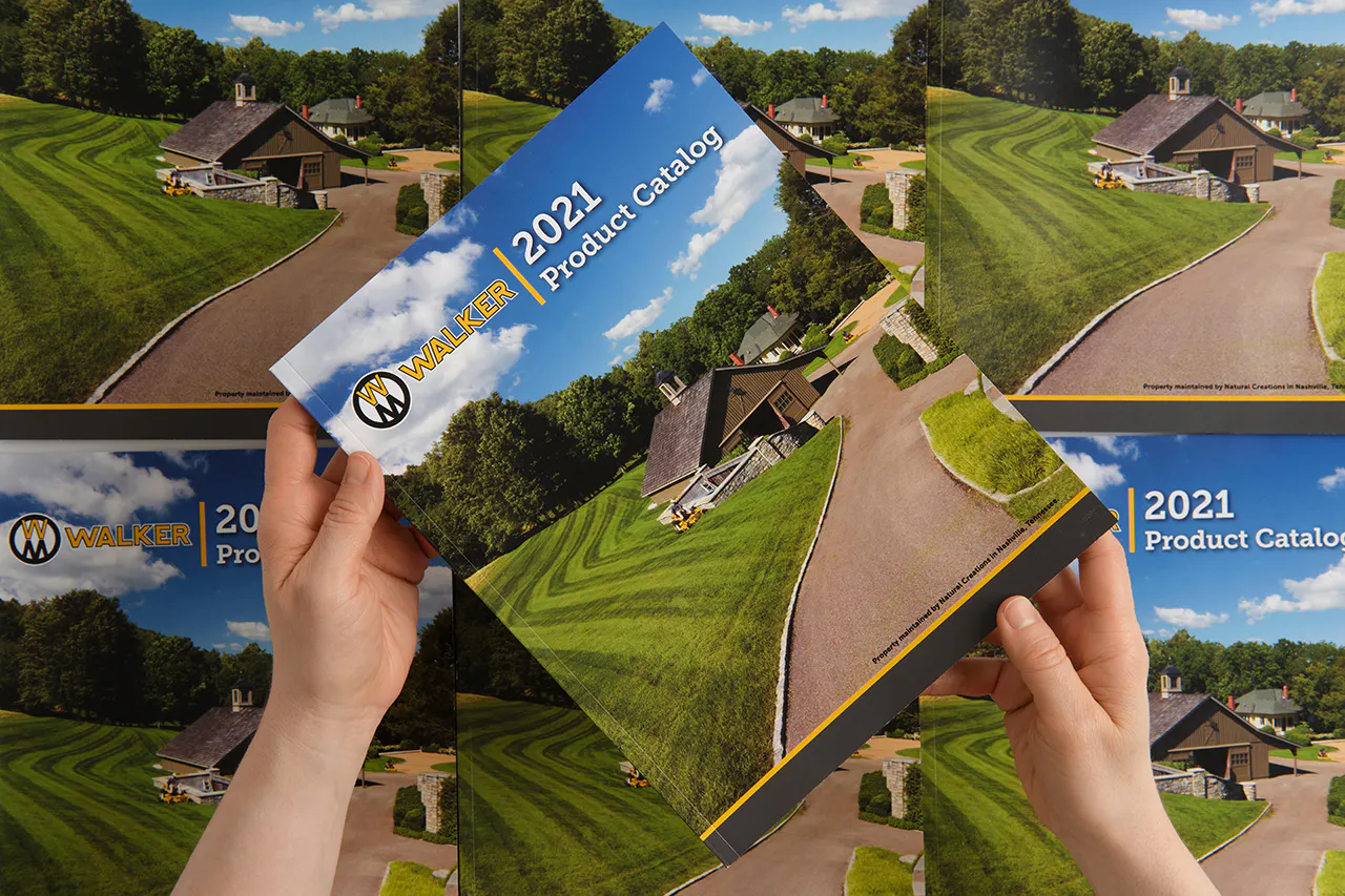 Two hands holding a Walker Mower product catalog printed with a house and a large green lawn on the cover.