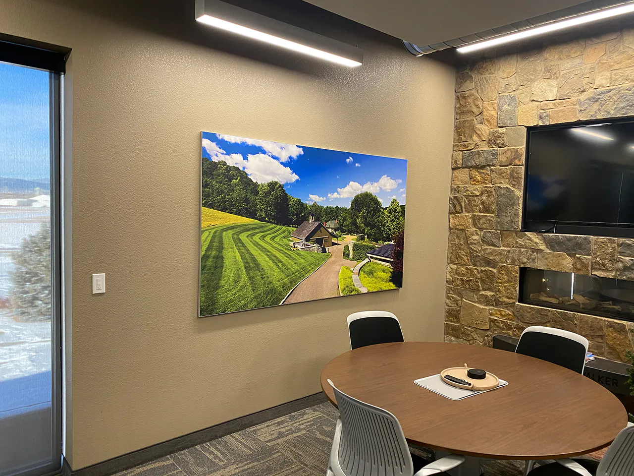 A meeting room with a round table and chairs in the middle and a picture of a house with a big green lawn on the wall.