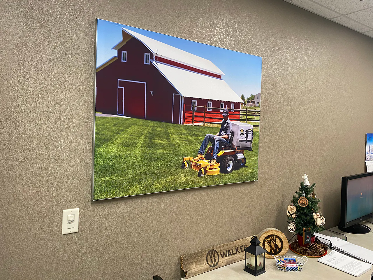 A picture hanging on a wall showing a big red barn and a man on a riding lawn mower over green grass.