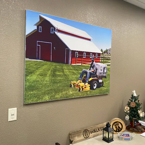 A picture hanging on a wall showing a big red barn and a man on a riding lawn mower over green grass.