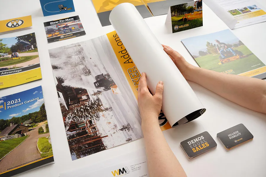 Two hands hold a rolled-up poster among various print marketing pieces, like business cards, table tents and catalogs.