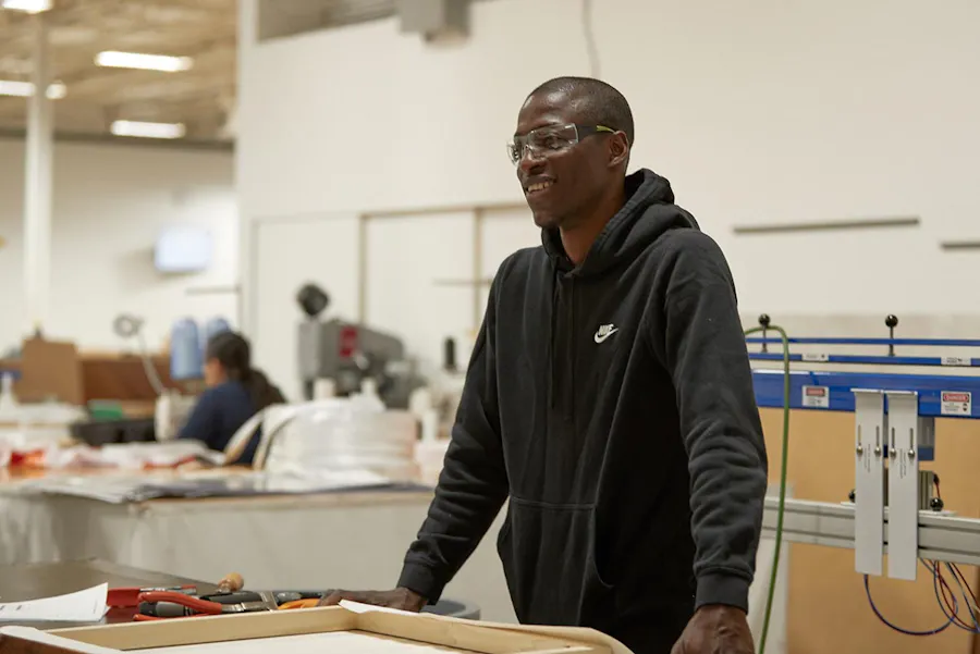 A man wearing safety glasses and a black hoodie standing at a table in a production facility.
