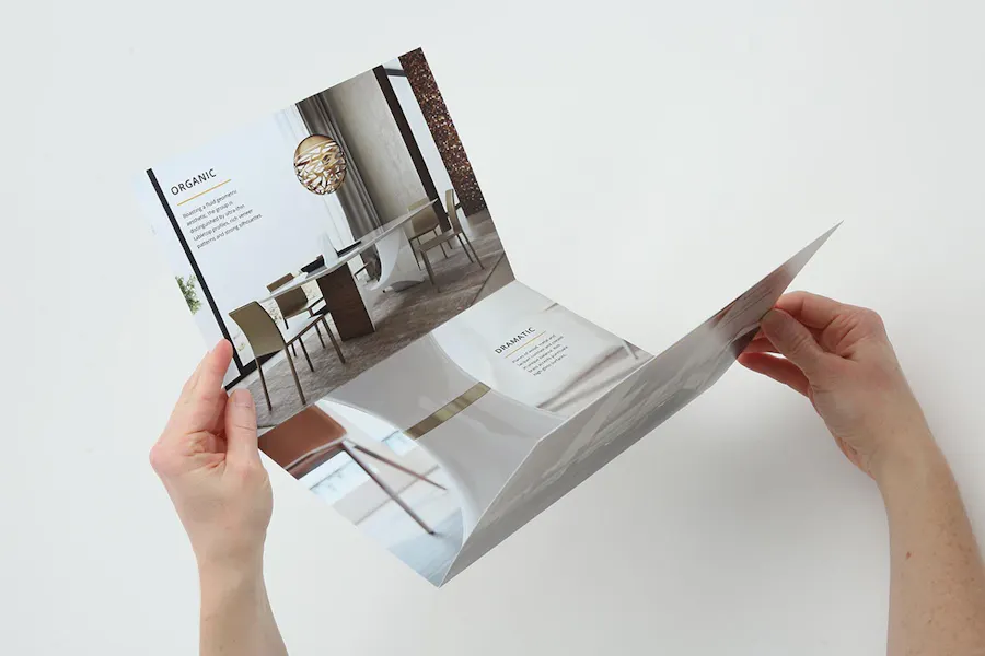 Two hands holding and unfolded tri-fold brochure with real estate imagery and marketing text.
