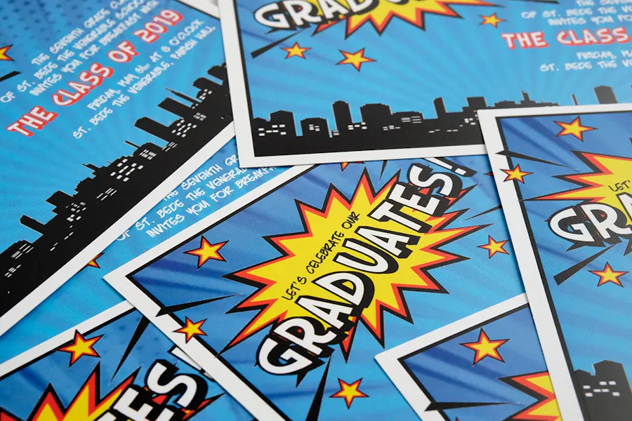A scattered pile of graduation invitations with a superhero design in blue, yellow and red.