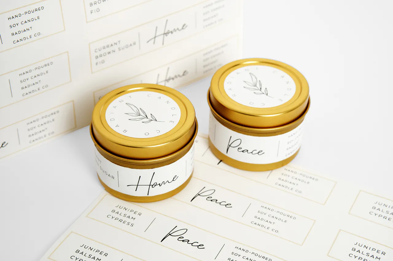 Two candles in gold tins with white labels printed with Radiant Candle Co. sitting on more sheets of labels.