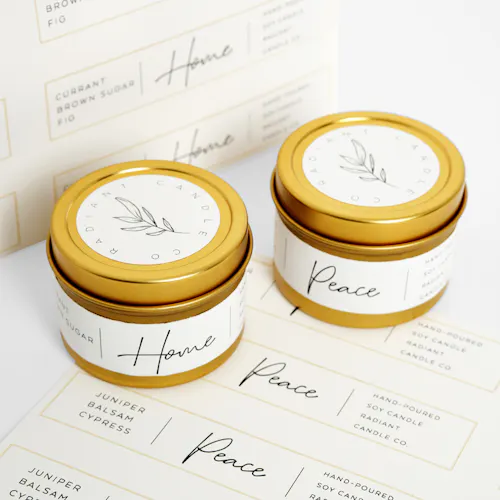 Two candles in gold tins with white labels printed with Radiant Candle Co. sitting on more sheets of labels.
