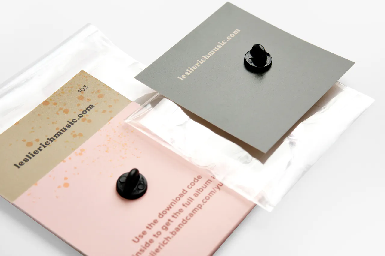 The backs of two enamel pins on custom backers printed with leslierichmusic.com.