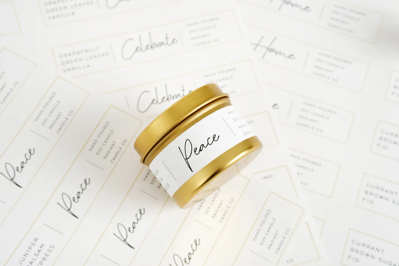 A candle in a gold jar and a white label laying on its side on top of sheets of labels printed with Peace and Home.