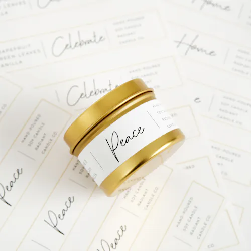 A candle in a gold jar and a white label laying on its side on top of sheets of labels printed with Peace and Home.