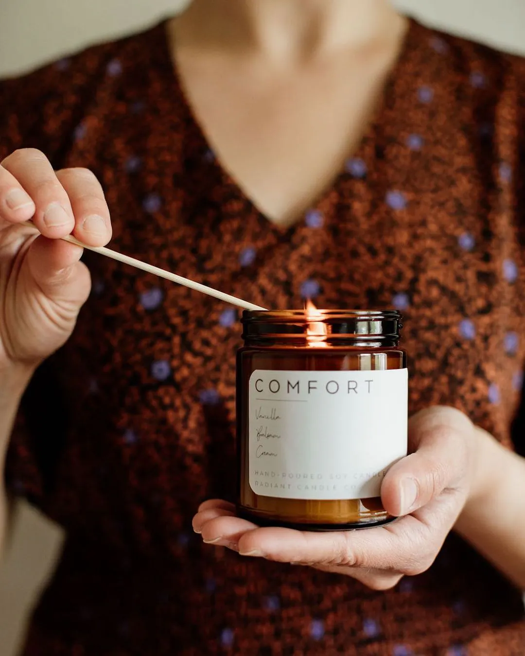 A woman wearing a brown dress with purple flowers and lighting a candle in a jar with a white label printed with Comfort.