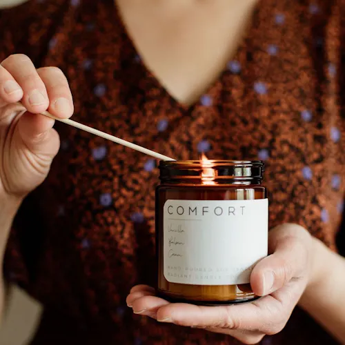 A woman wearing a brown dress with purple flowers and lighting a candle in a jar with a white label printed with Comfort.