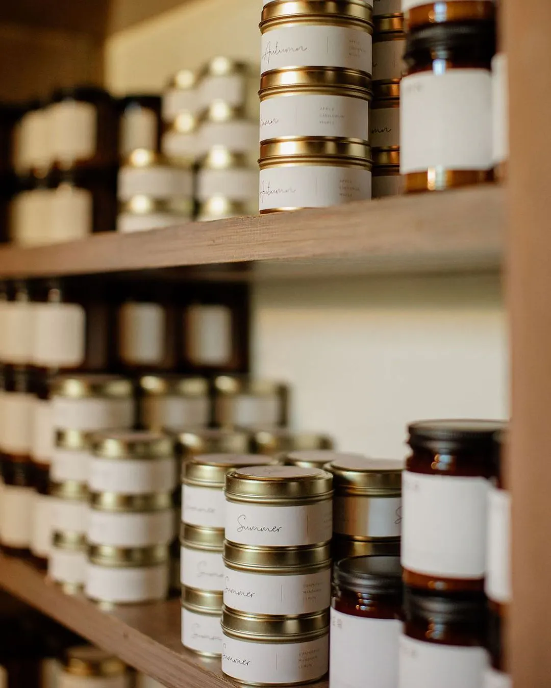 Stacks of candles in jars and tins on wooden shelves with custom white labels.