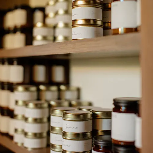 Stacks of candles in jars and tins on wooden shelves with custom white labels.