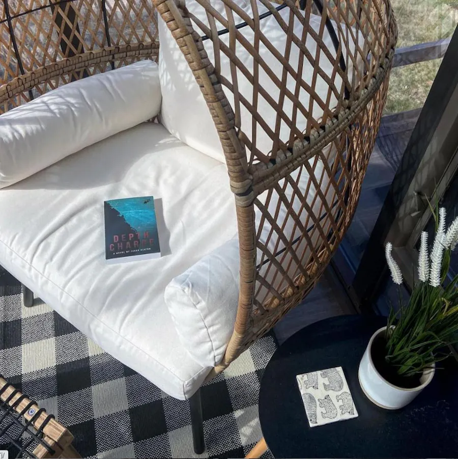 A paperback book on a wicker chair on a black and white checkered rug next to a black side table with a plant on top.