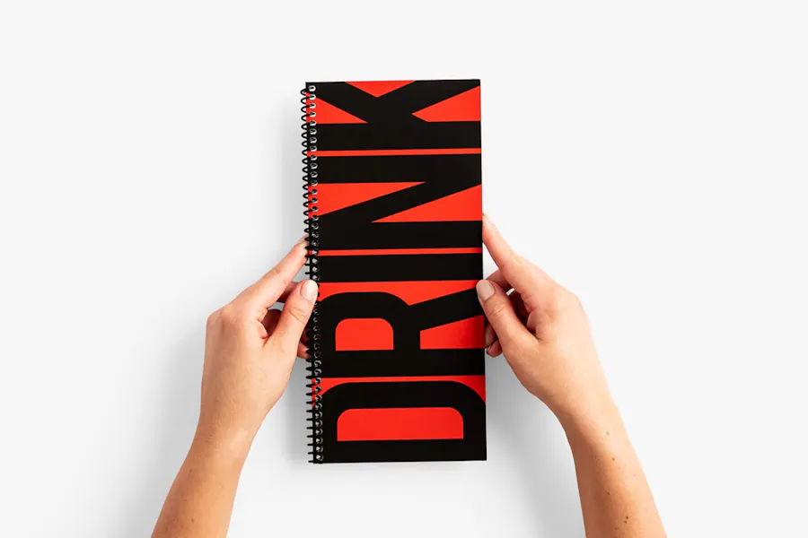 Two hands holding a spiral bound drink menu printed with a red background and DRINK in black text.