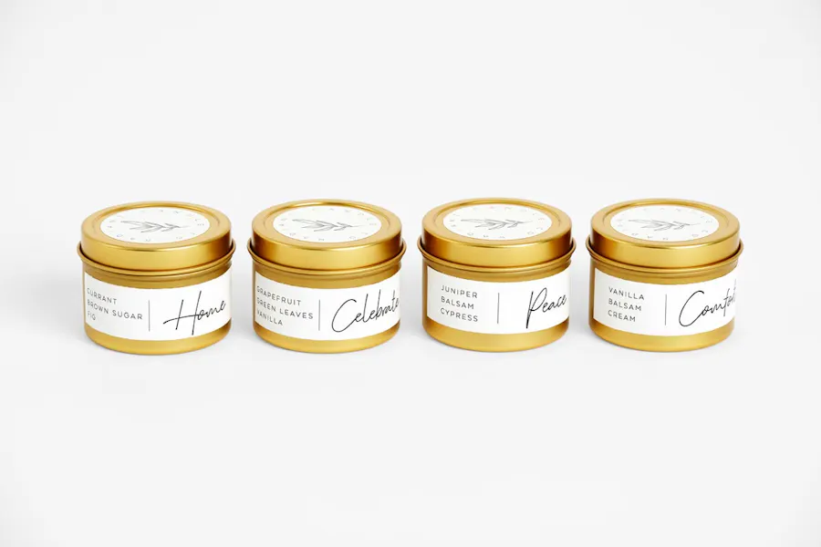 Four candles in gold tins with custom packaging labels on them in white with black cursive writing.
