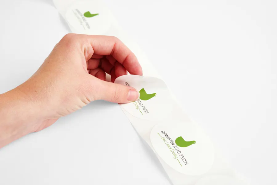A hand peeling an oval-shaped product label off its backing, printed with Farmington Road Fresh.