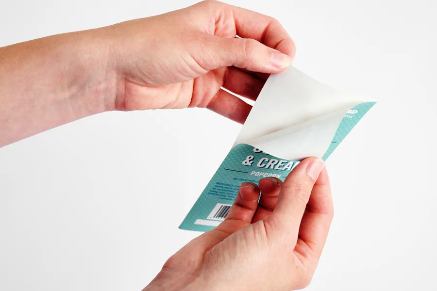 Two hands peeling a teal popcorn label off of its white crack n peel backing stock.