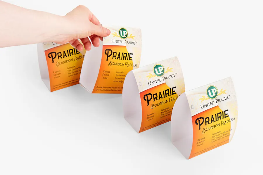A hand lifting up one custom table tent from a line of table tents with a Prairie Bourbon Radler recipe on it.