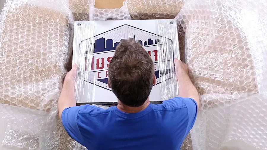 A man in a blue T-shirt putting custom aluminum signs into a packaging box with bubble wrap.