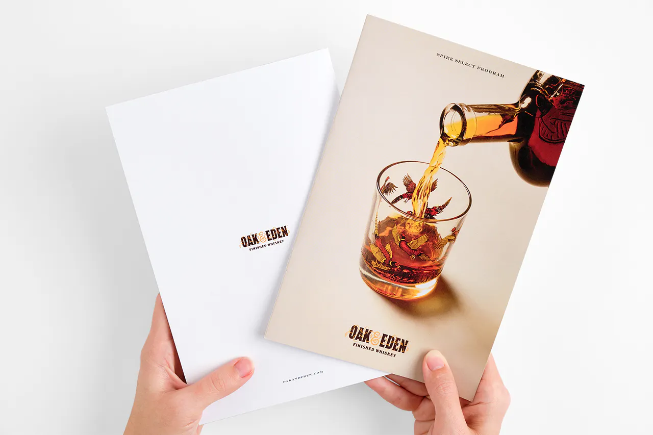 Two hands holding two Oak & Eden marketing booklets with a glass of whiskey on one cover.
