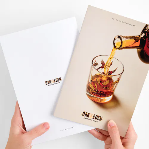 Two hands holding two Oak & Eden marketing booklets with a glass of whiskey on one cover.