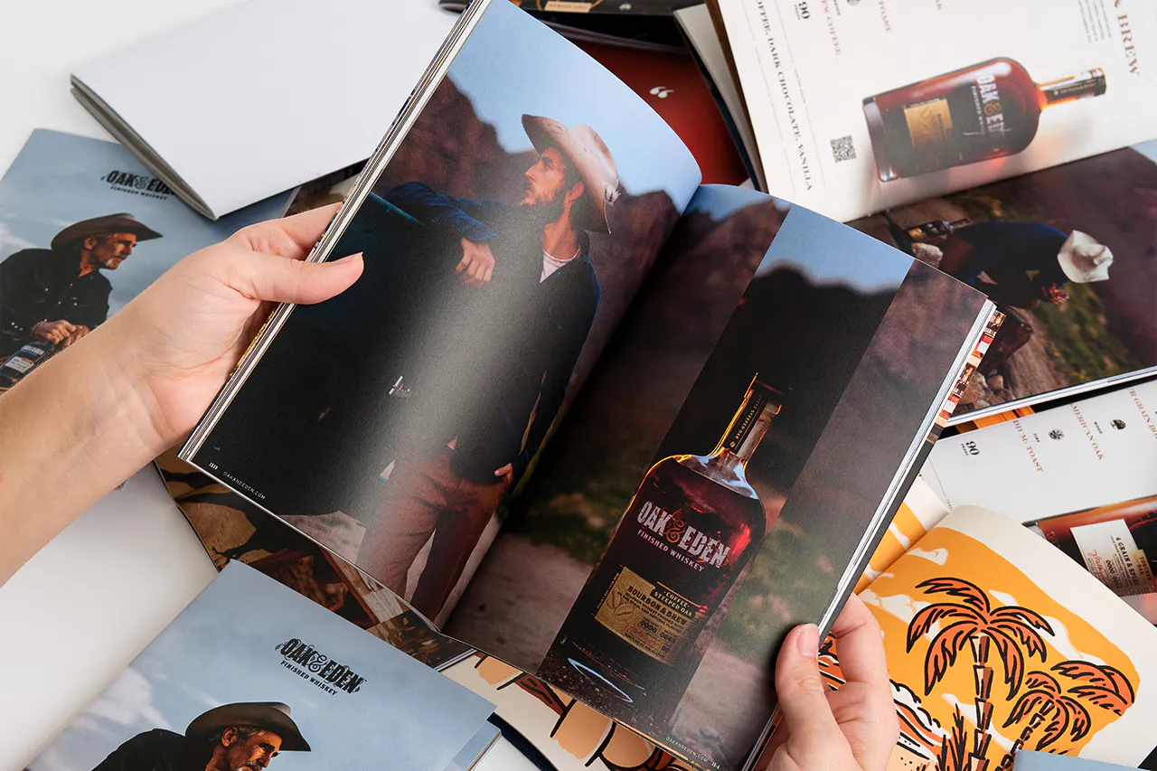 Two hands flipping through an Oak & Eden brand book with images of a man wearing a cowboy hat and a whiskey bottle.