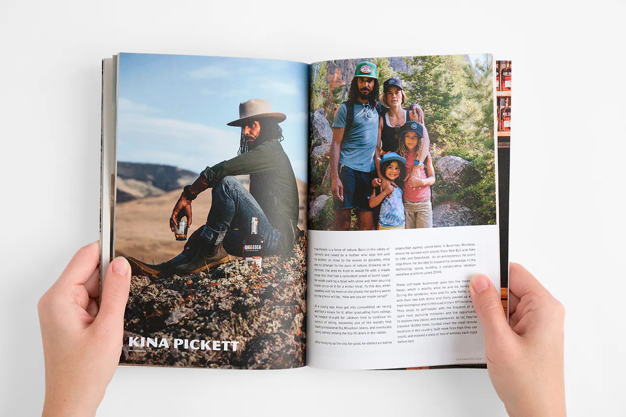 Two hands holding open a brand book with images of a man sitting on a rock holding a glass of whiskey and a family hiking.