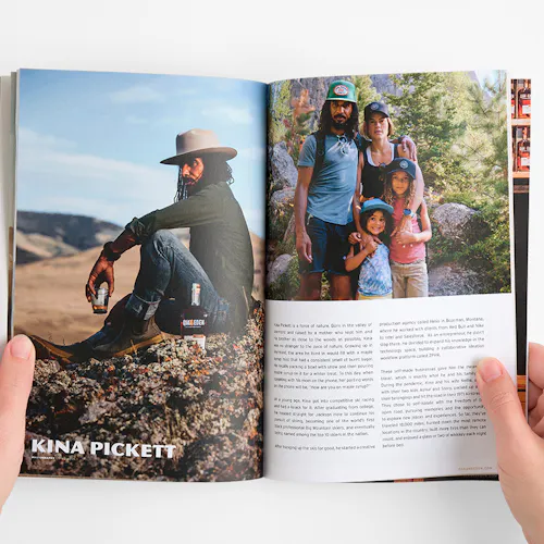 Two hands holding open a brand book with images of a man sitting on a rock holding a glass of whiskey and a family hiking.