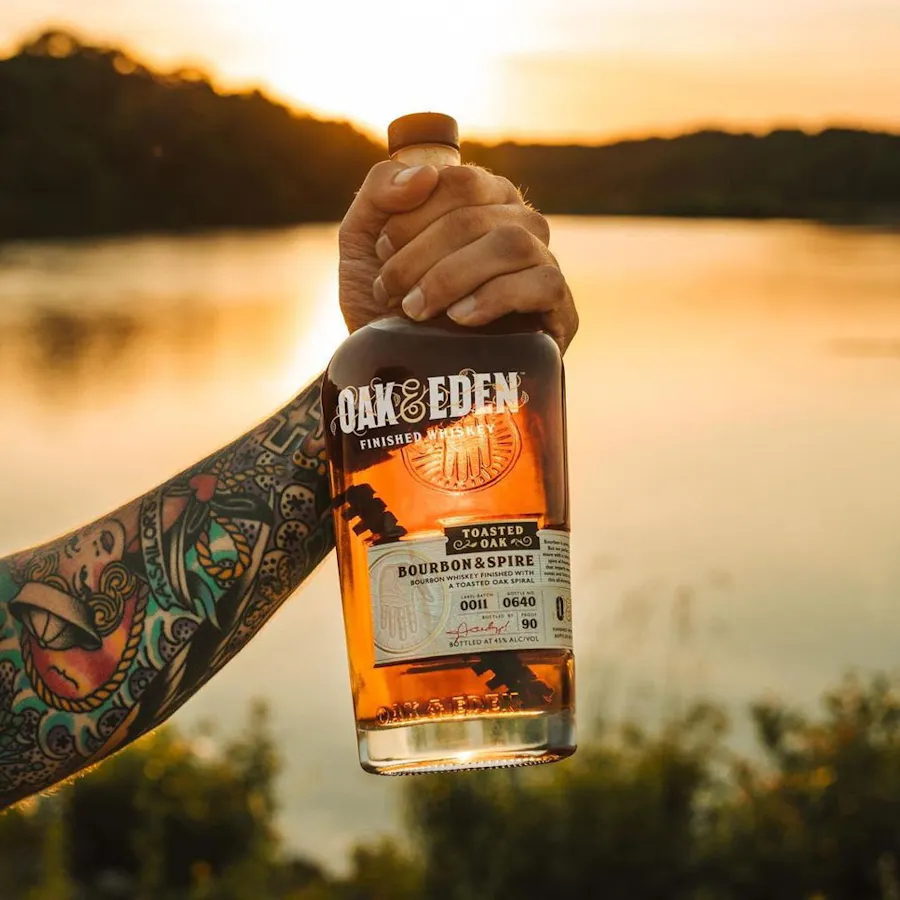 A hand with tattoos up the forearm holding a bottle of Oak & Eden whiskey by a lake at sunset.