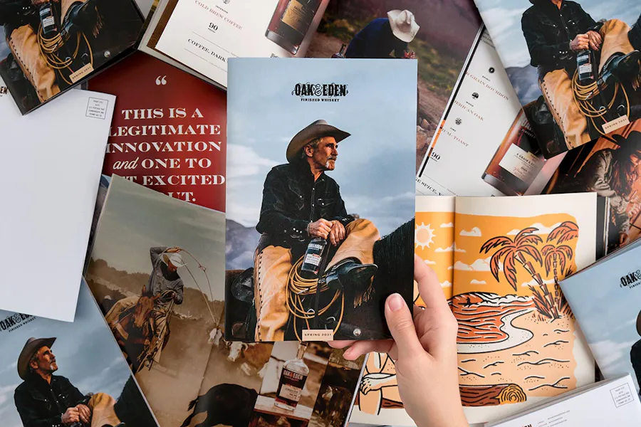 A hand holding a direct mail booklet with "Oak & Eden" and a cowboy on the front above other marketing materials.