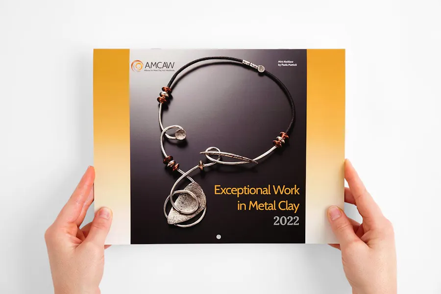 Two hands holding a custom printed calendar with a saddle stitch binding and a metalwork necklace on the cover.