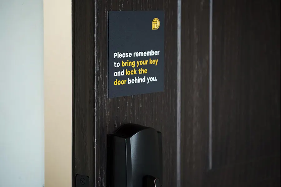 A sign above a door handle with Please remember to bring your key and lock the door behind you. in white and yellow.