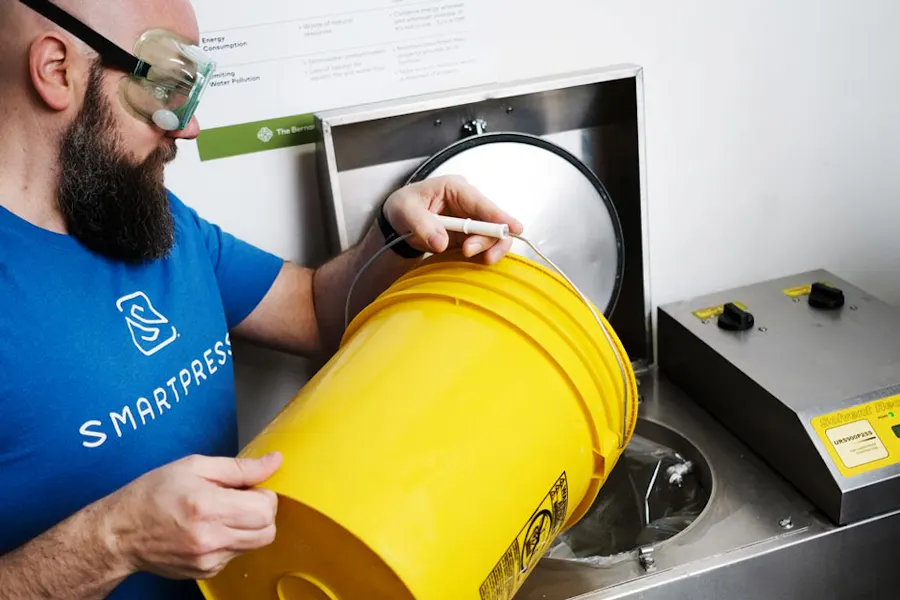 A man in a blue Smartpress T-shirt wearing safety goggles and pouring contents of a yellow pail into a solvent machine.