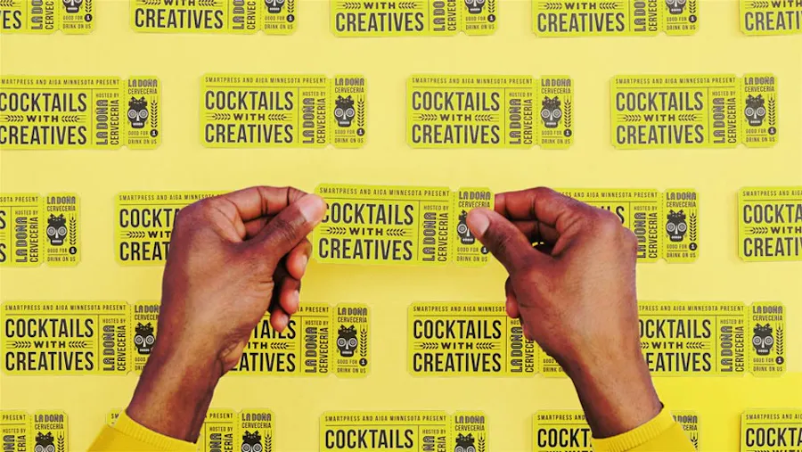 Two hands holding a custom drink ticket printed in yellow and black with more tickets around it.