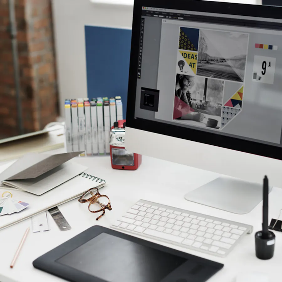 A desktop computer showing a graphic design program with a keyboard, glasses, notebooks and pens around it.