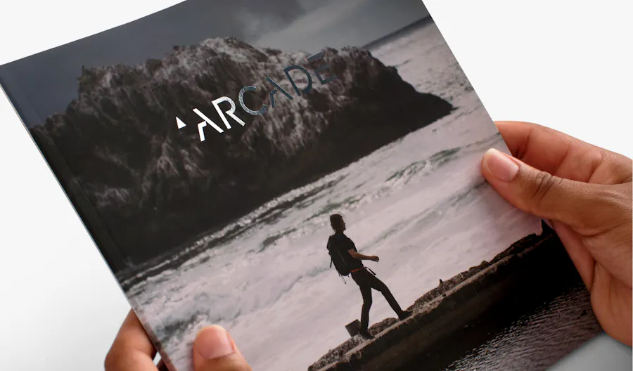 Two hands holding a marketing booklet printed printed with Arcade in silver foil and a person hiking by water on the cover.